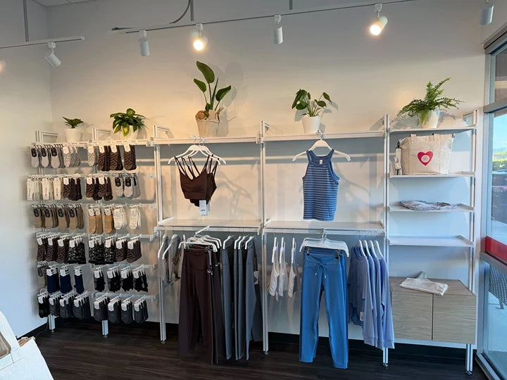A retail clothing display featuring multiple white metal racks holding various apparel items including tops, pants, and a large stack of socks on the left. The store has a clean white wall, overhead track lighting, and small potted plants on the shelves.
